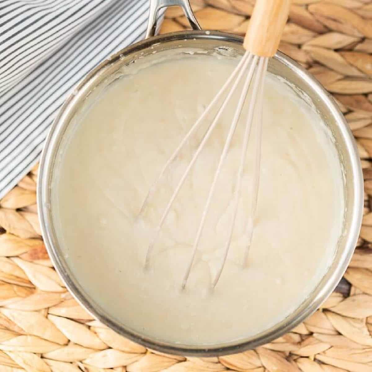 A metal saucepan filled with creamy white sauce is being whisked with a metal whisk—a perfect step if you're learning how to make a roux. The pan rests on a woven placemat next to a striped cloth.