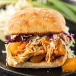 A close-up of a Hawaiian BBQ Chicken Sandwich on a ciabatta roll, filled with shredded chicken, pineapple chunks, and colorful shredded cabbage on a black plate.