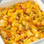 A close-up of a keto loaded cauliflower casserole topped with melted cheese, crispy bacon bits, and chopped green onions in a white square baking dish.