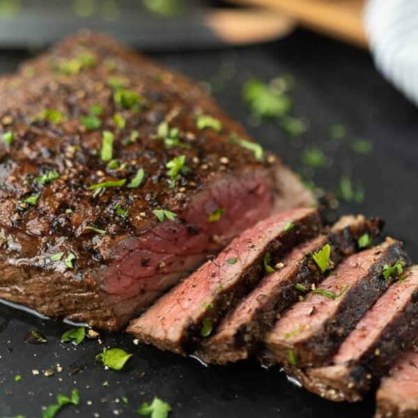 Close-up of a juicy, medium-rare roast beef, seasoned with herbs, pepper, and a hint of London Broil Marinade, partially sliced on a dark surface. Fresh parsley is sprinkled on top, adding a touch of green to the dish.