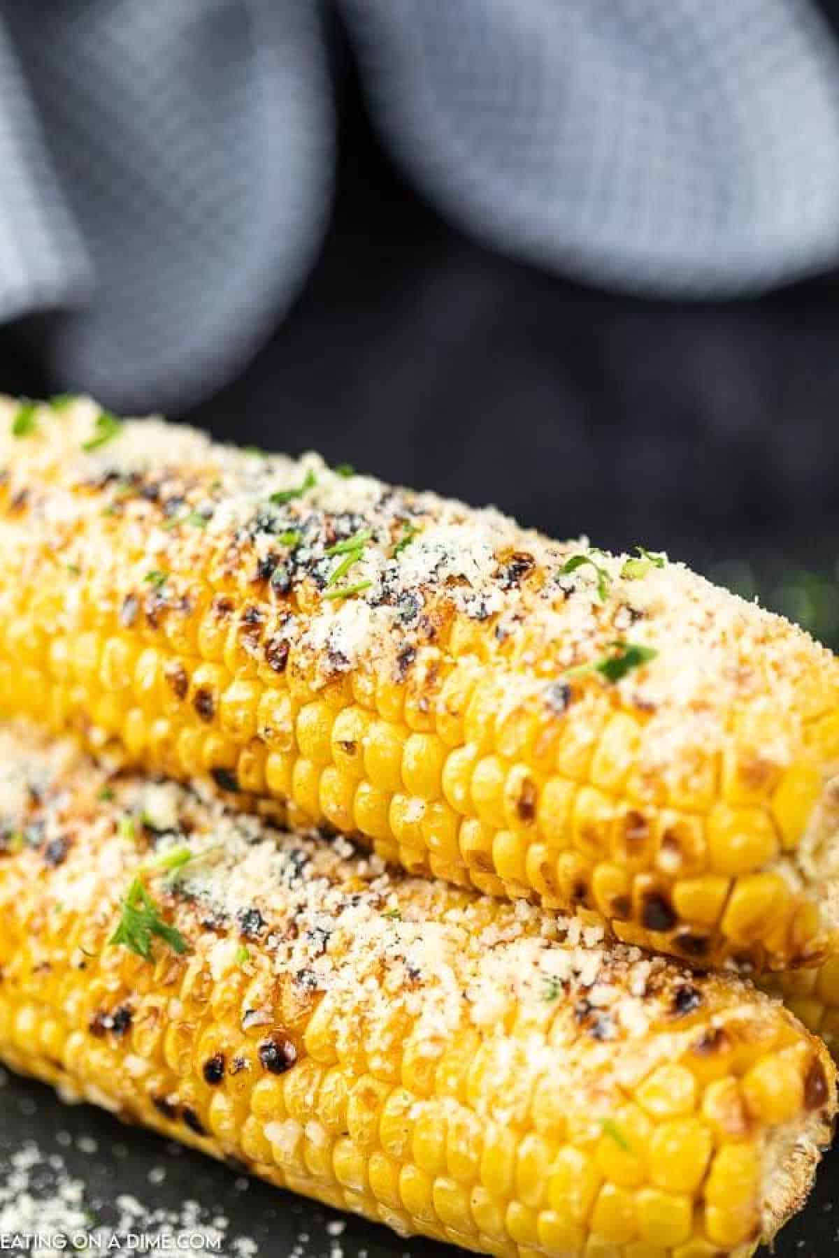 Two Parmesan grilled corn on the cob sprinkled with grated cheese and garnished with chopped herbs. The corn has a slightly charred appearance, and the background is blurred.