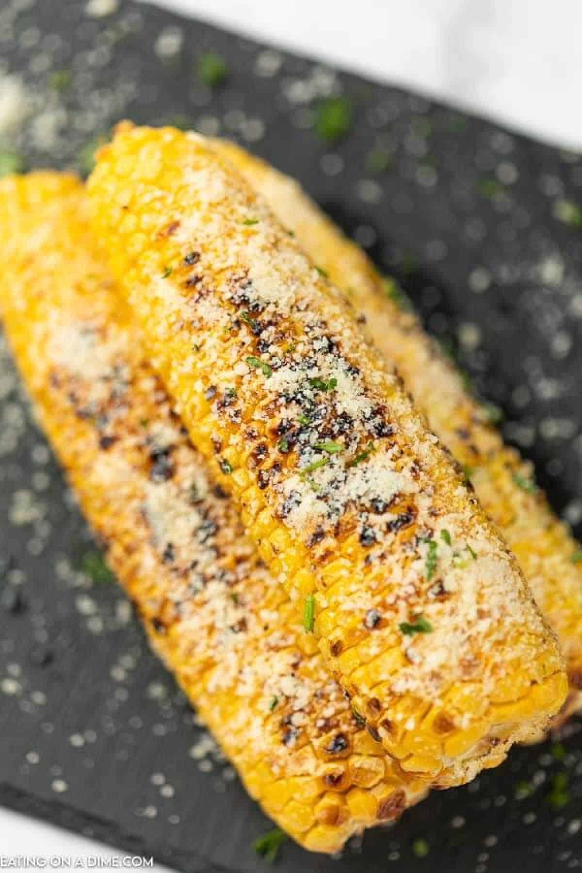Three Parmesan Grilled Corn cobs are stacked on a dark surface, topped with grated cheese and sprinkled with herbs. They showcase char marks and a golden appearance, making them irresistible.