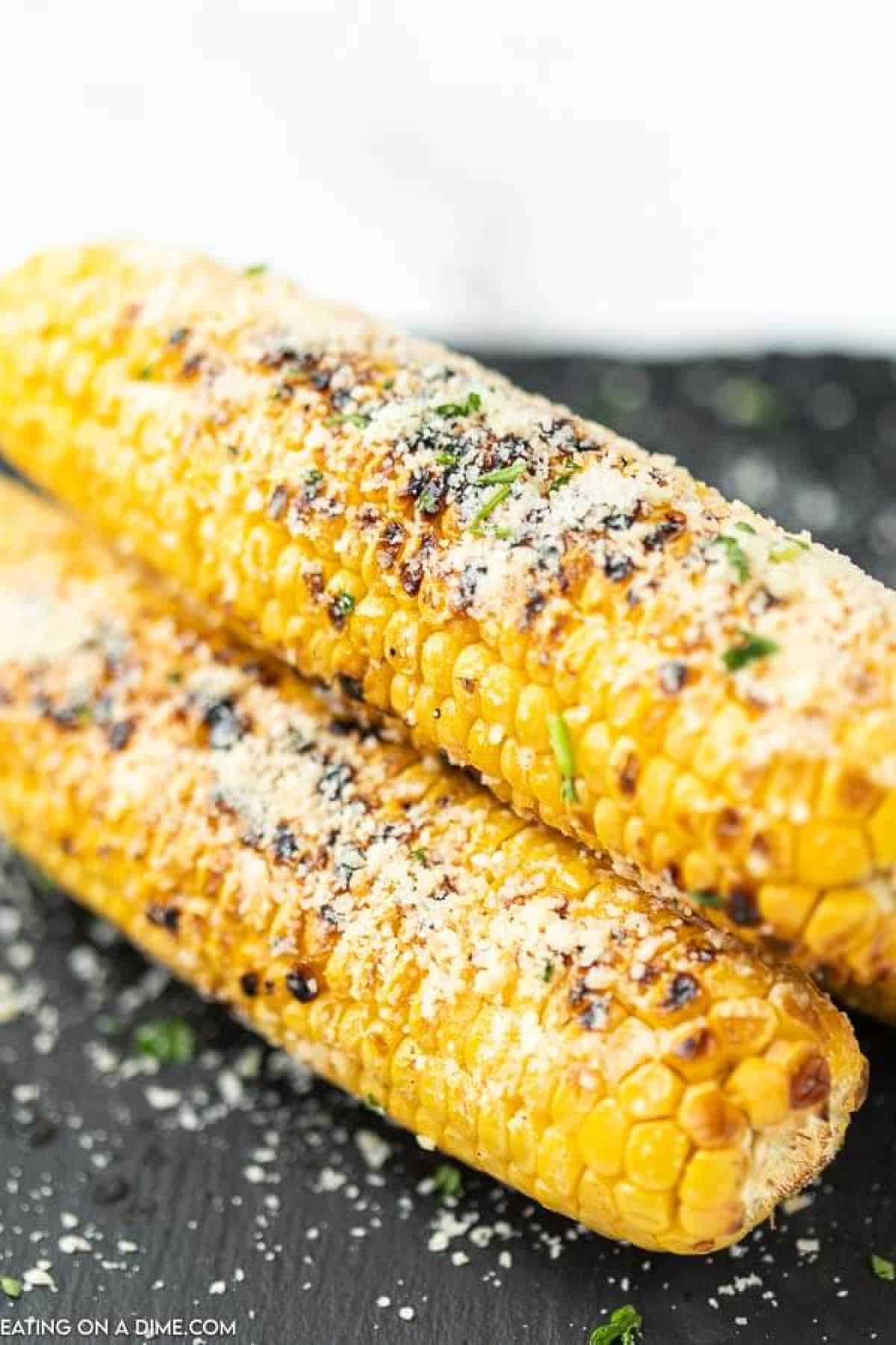Parmesan grilled corn on the cob is the perfect combination of butter, garlic and parmesan cheese. Enjoy this easy and tasty side dish. Two grilled corn on the cob are topped with grated Parmesan cheese and sprinkled with herbs, resting on a dark surface. The Parmesan grilled corn features charred grill marks, adding texture and a golden color.