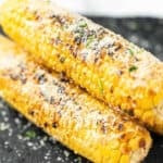 Two Parmesan Grilled Corn on the Cob, topped with grated cheese and sprinkled herbs, rest on a dark surface. Char marks and a golden-yellow color make this dish irresistibly appetizing.