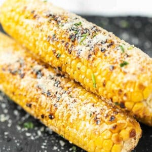 Two Parmesan Grilled Corn on the Cob, topped with grated cheese and sprinkled herbs, rest on a dark surface. Char marks and a golden-yellow color make this dish irresistibly appetizing.