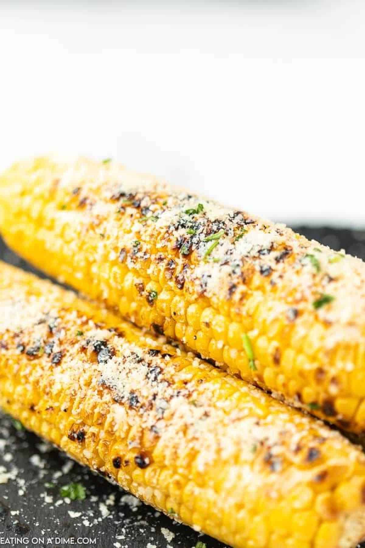 Two ears of Parmesan grilled corn on the cob are topped with grated cheese and herbs, set on a dark surface. They appear golden and slightly charred, with a sprinkling of cheese enhancing their texture.