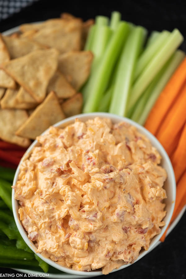 pimento cheese in white bowl on platter with vegetables and pita chips.
