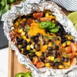 A Santa Fe Chicken foil pack filled with baked chicken, melted cheddar cheese, black beans, corn, cherry tomatoes, jalapeño slices, and cilantro is displayed on a wooden board with lime wedges nearby.