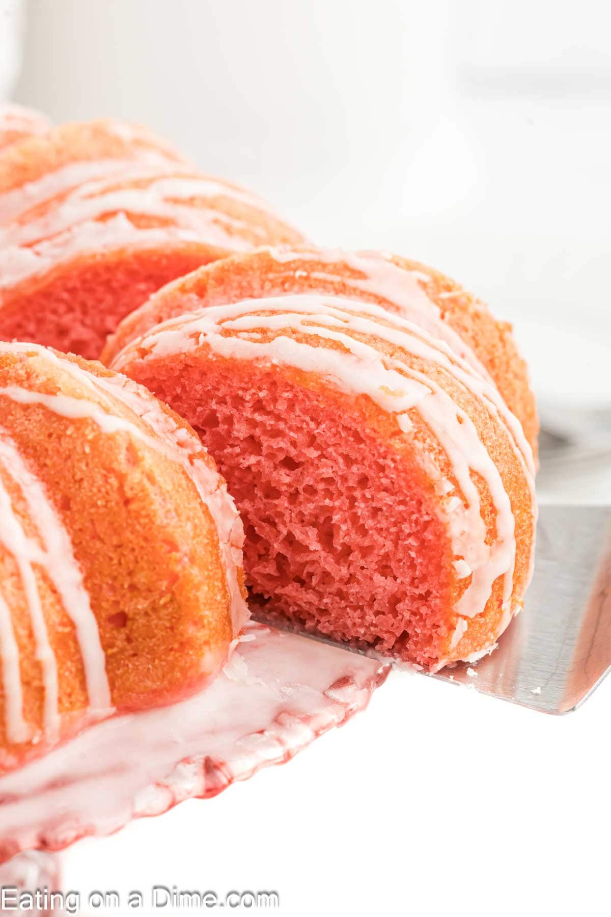 Strawberry Bundt Cake on a cake platter drizzled with white glaze with a slice cut and on a spatula
