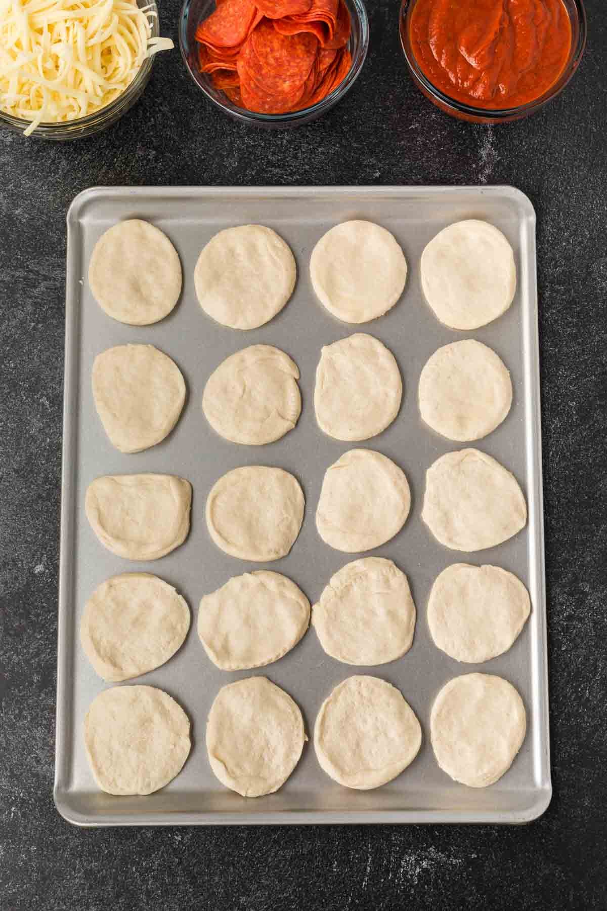 A baking sheet with 20 round pieces of raw dough, ready for a biscuit pizza recipe. Above the tray are bowls of shredded cheese, pepperoni slices, and pizza sauce on a dark countertop.