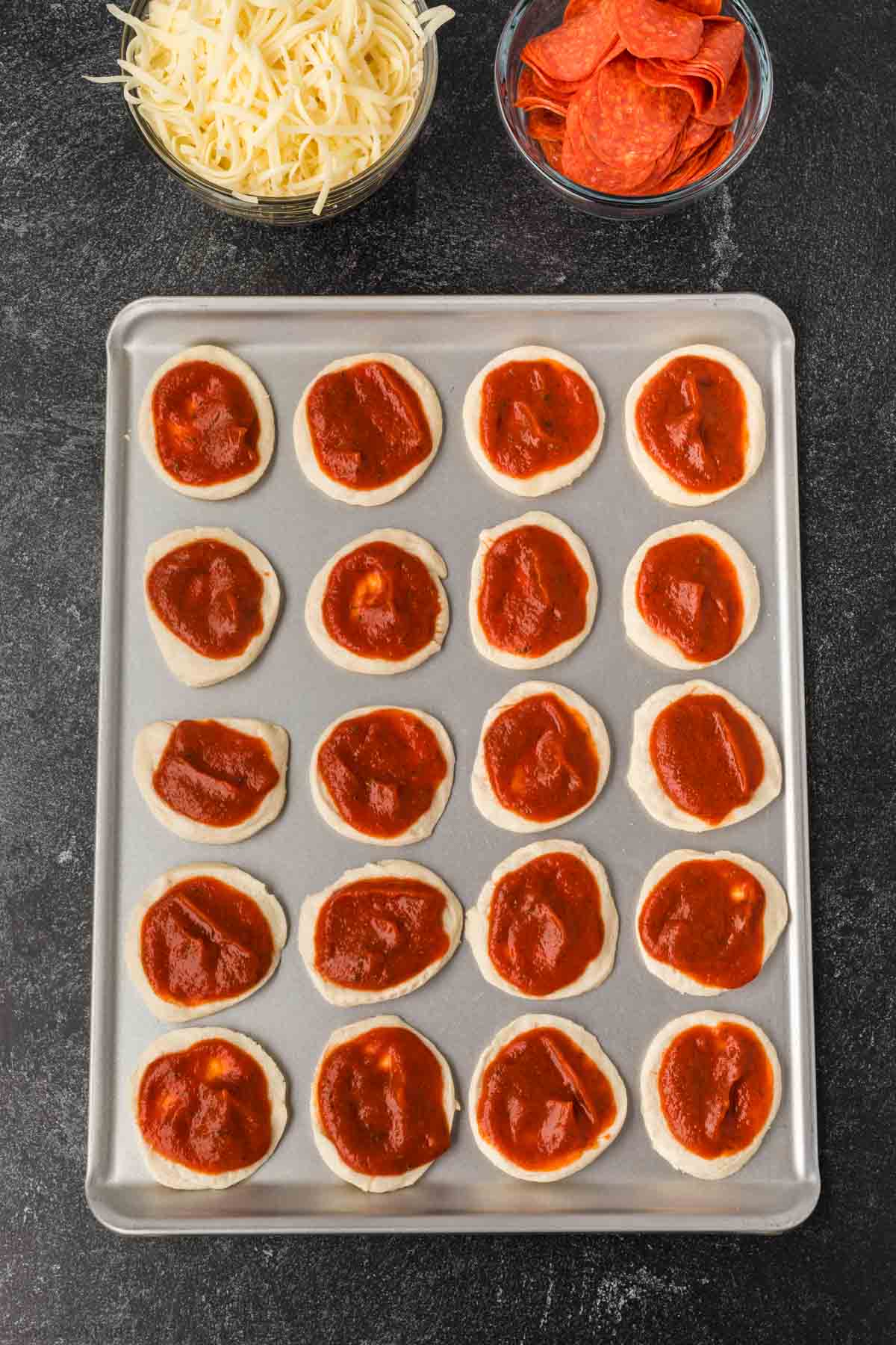 A baking sheet with rows of mini biscuit pizza recipe dough rounds topped with tomato sauce, next to bowls of shredded cheese and pepperoni slices on a black countertop.