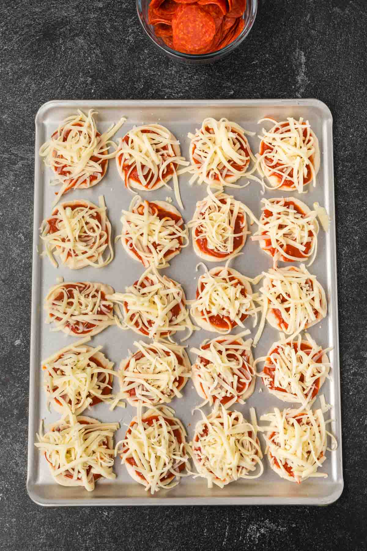 A baking sheet filled with mini biscuit pizzas topped with shredded cheese and tomato sauce, ready to be baked. A bowl of pepperoni slices is visible at the top—perfect for trying a fun biscuit pizza recipe.