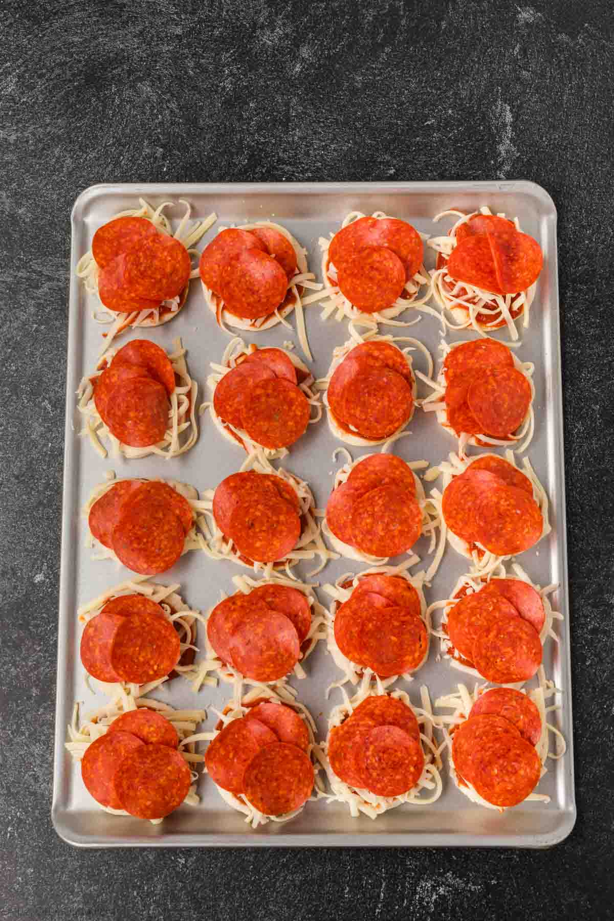A baking sheet filled with rows of uncooked mini pizzas, made using a biscuit pizza recipe, each topped with shredded cheese and slices of pepperoni, set on a dark countertop.
