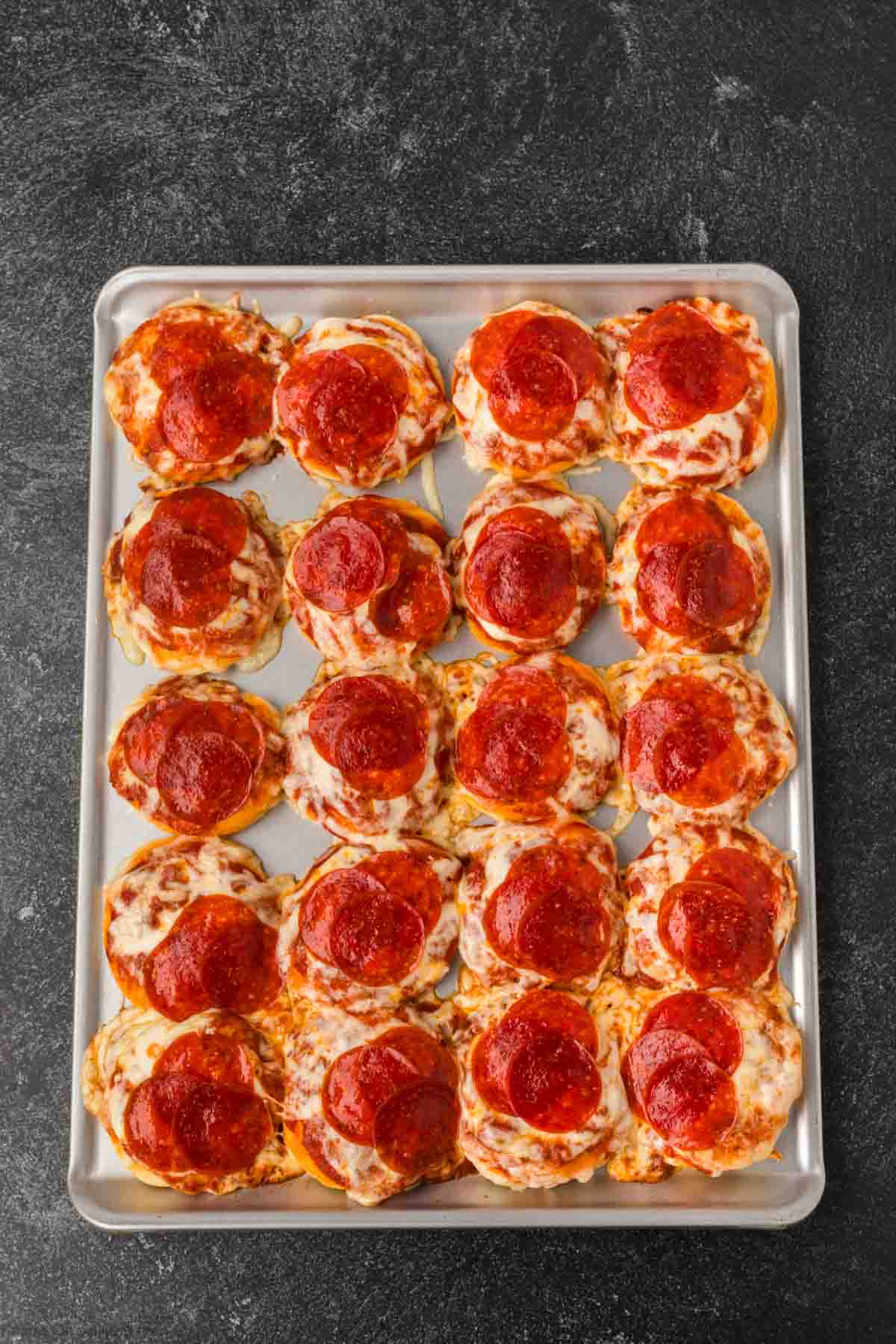 A baking sheet filled with twenty mini biscuit pizza recipe creations, each topped with melted cheese and slices of pepperoni, on a dark textured surface.