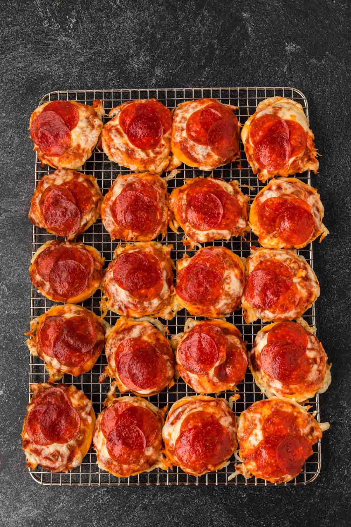 Mini pepperoni pizzas made with a biscuit pizza recipe line up on a wire cooling rack, topped with melted cheese and pepperoni slices, set against a dark textured surface.