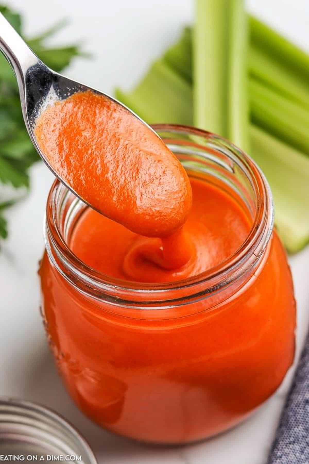 A spoon scooping out a serving of buffalo sauce from a jar of homemade buffalo sauce with celery sticks behind it.  