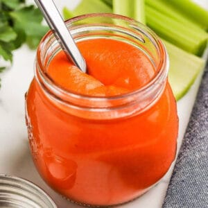 A close-up photo of a jar of homemade buffalo sauce with sticks of celery and parsley behind it.