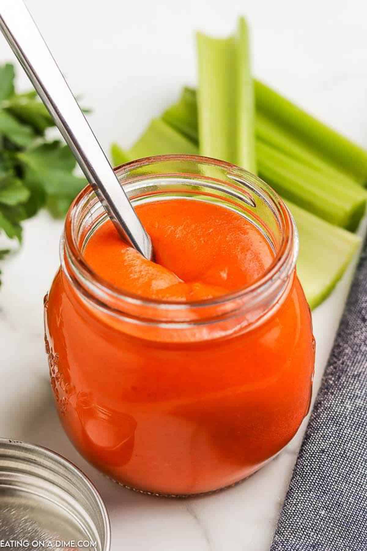 A jar of homemade buffalo sauce with a spoon in the buffalo sauce and with sticks of celery and parsley behind it.  