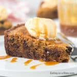 A slice of crock pot chocolate chip cookie pie topped with a scoop of vanilla ice cream and drizzled with caramel sauce sits on a white plate with a fork.
