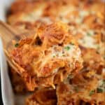 A wooden spoon lifts cheesy baked ziti from a chicken nugget casserole, topped with chopped herbs and a softly blurred green background.