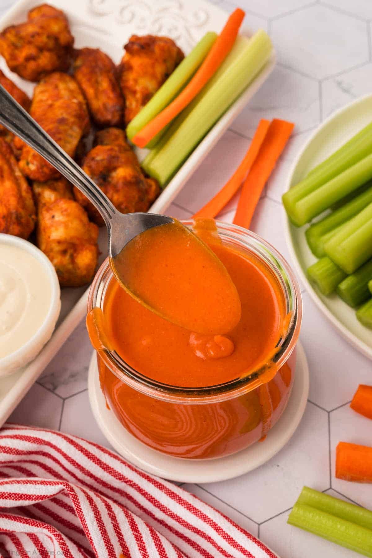 Buffalo Sauce in a clear jar with a serving on a spoon with platter of buffalo wings and carrots and celery on the side