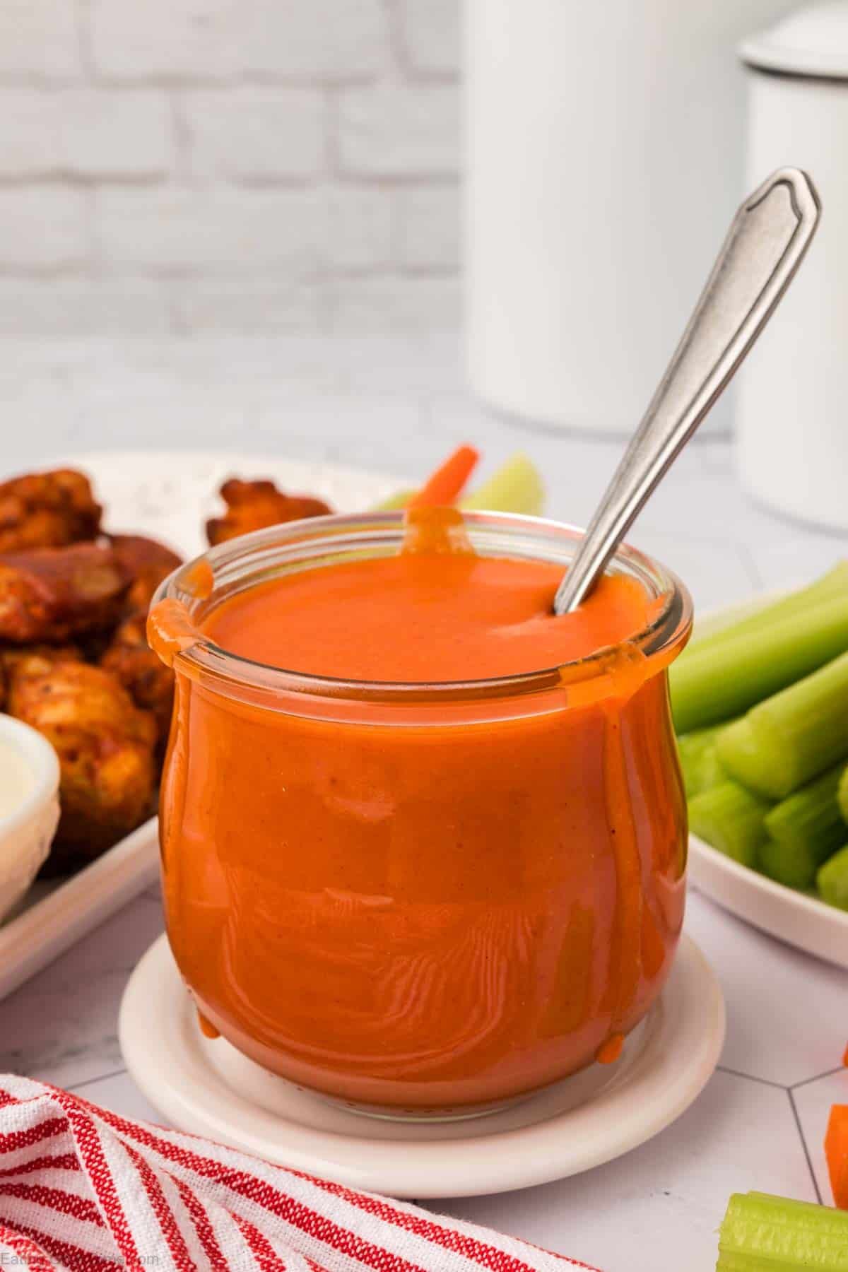 Buffalo Sauce in a clear jar with a spoon and a platter of fresh celery and buffalo wings in the background