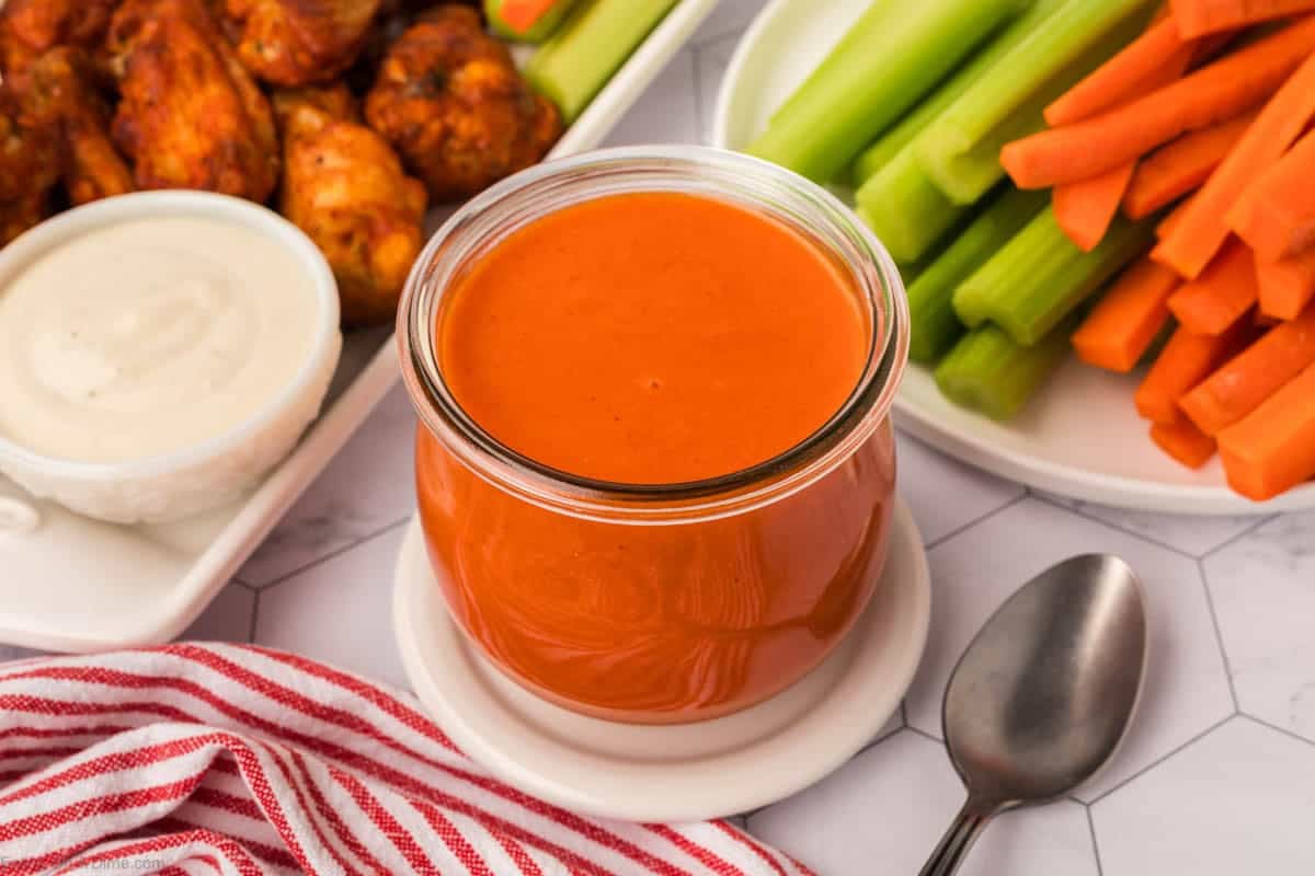 Buffalo Sauce in a clear jar with a platter of buffalo wings, celery and carrots on the side