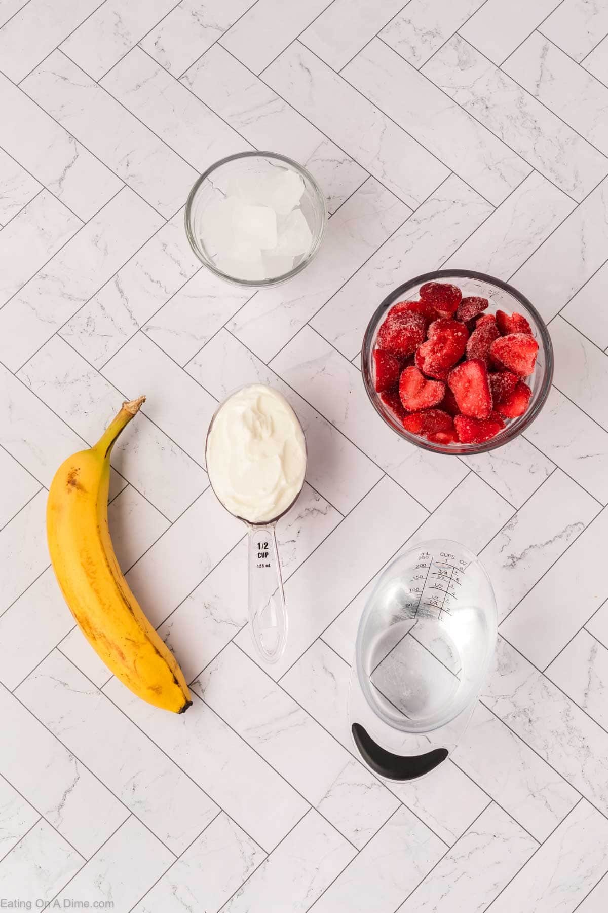 A banana, a measuring cup of yogurt, ice cubes, frozen strawberries, and water are arranged on a white, herringbone-patterned countertop—perfect ingredients for a refreshing Strawberry Banana Yogurt Smoothie.