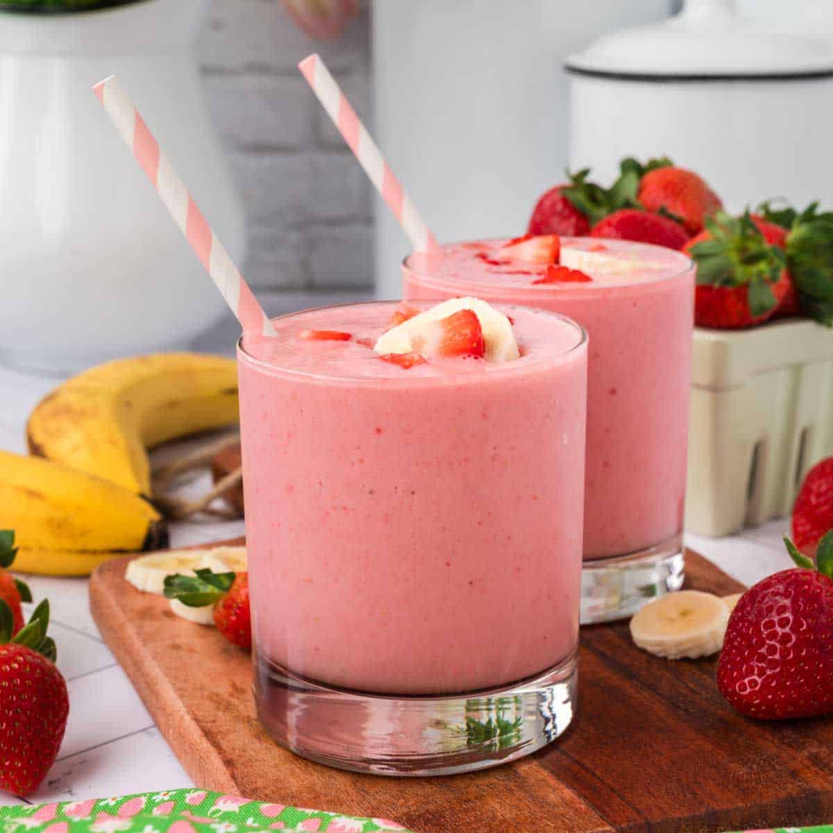 Two glasses of Strawberry Banana Yogurt Smoothie with striped straws, garnished with banana and strawberry slices, are on a wooden board surrounded by fresh strawberries and bananas.
