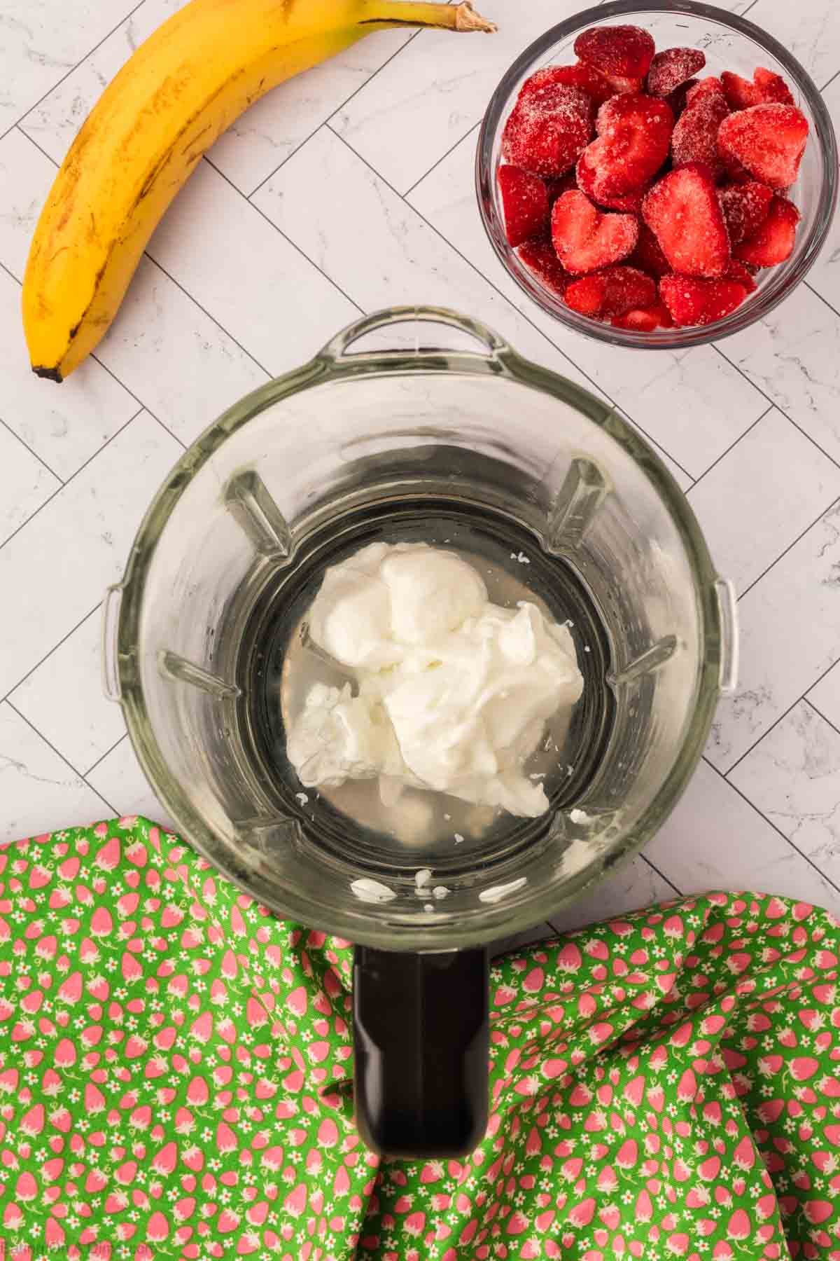 A blender with yogurt inside sits on a counter, ready to blend a delicious Strawberry Banana Yogurt Smoothie. Nearby are a banana, a bowl of frozen strawberries, and a green cloth with a pink floral pattern.