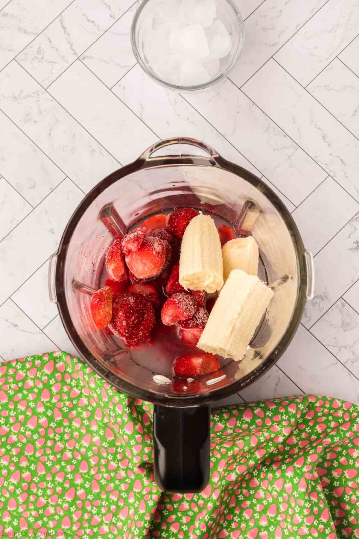 A blender filled with strawberries, banana, and liquid—ready to become a Strawberry Banana Yogurt Smoothie. Nearby, a glass of ice cubes and a green cloth with pink floral patterns rest on a white tiled surface.