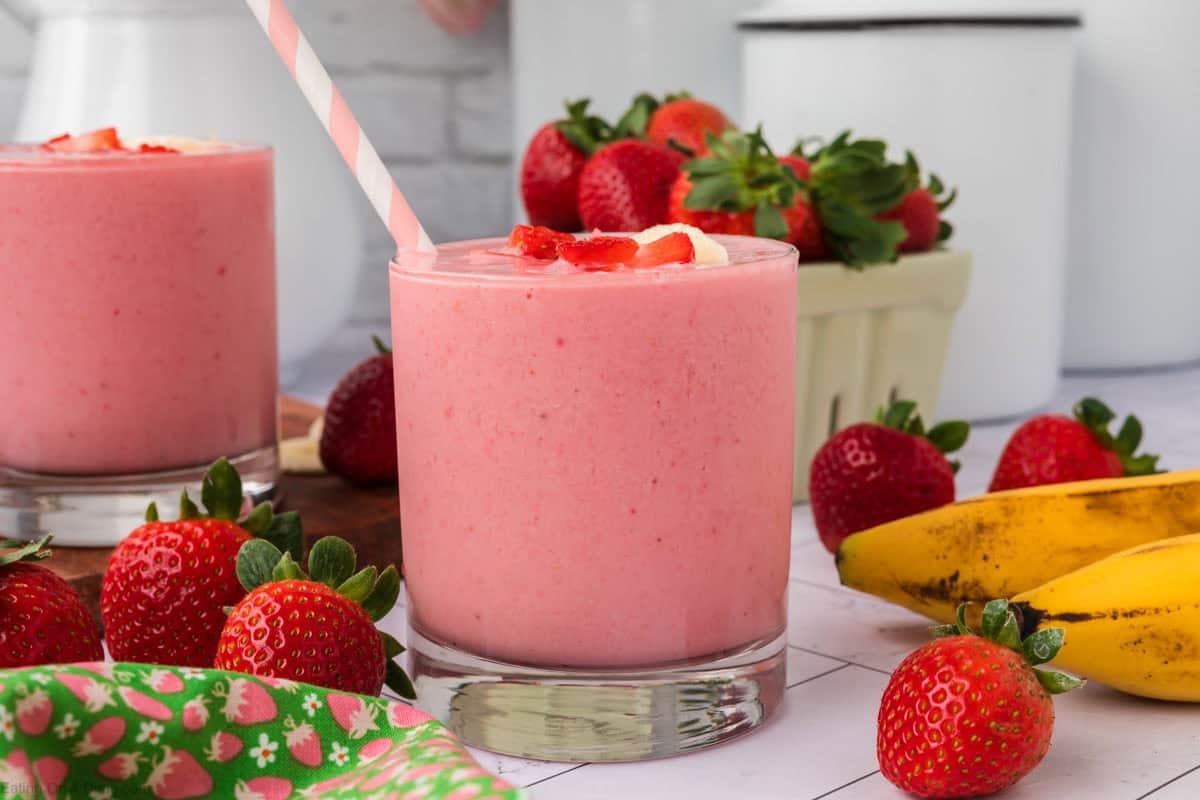 A glass of pink Strawberry Banana Yogurt Smoothie with a straw, topped with sliced strawberries and banana. Fresh strawberries and bananas are scattered around, with another smoothie in the background.