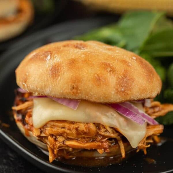 A pulled chicken sandwich on a ciabatta bun with melted cheese and sliced red onions on a black plate.
