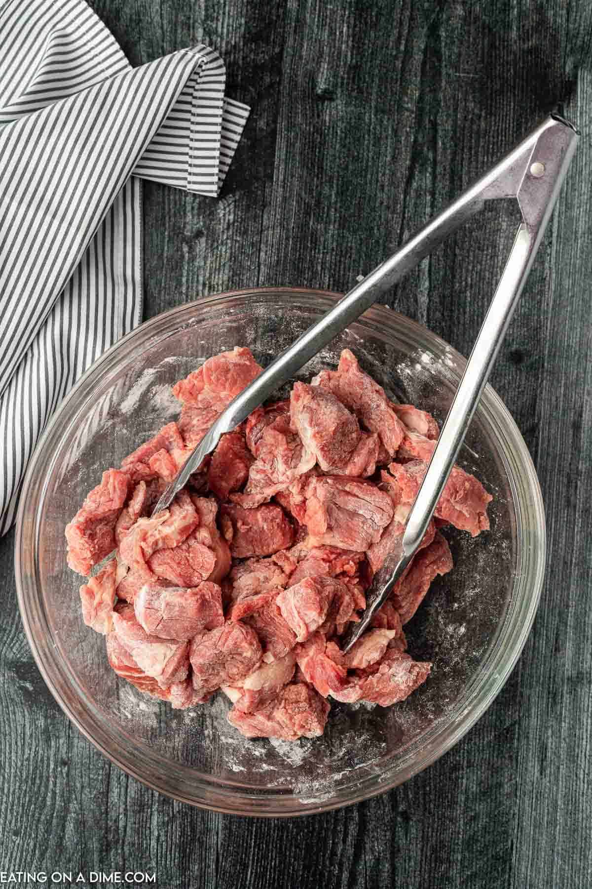 Mixing beef tips with flour in a bowl with tongs