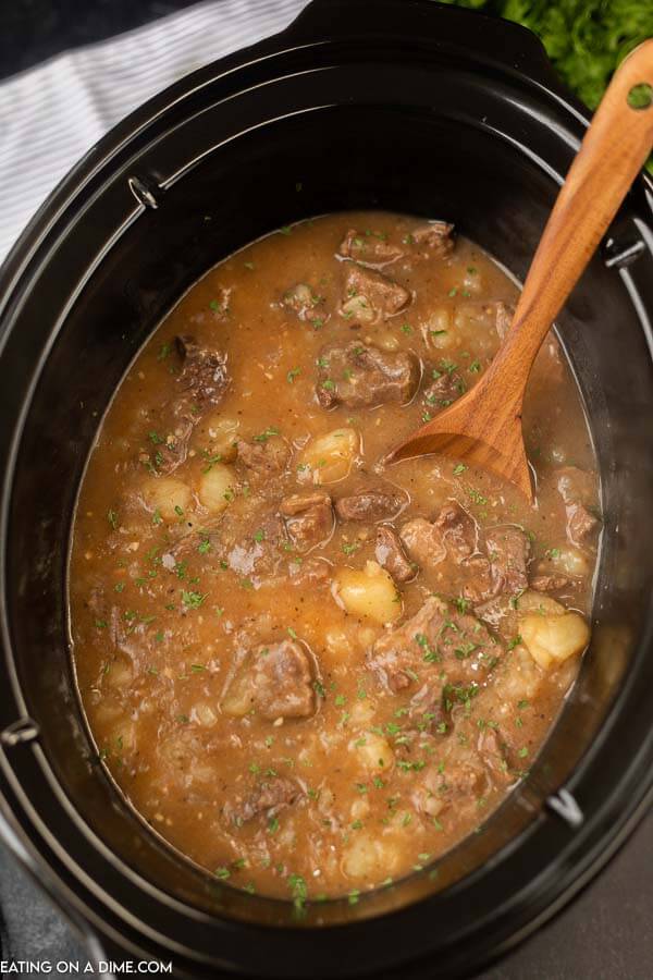 A black slow cooker of steak and potato stew with a wooden spoon in it.  
