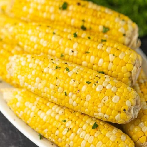 5 or 6 grilled corns on the cobs on a white plate topped with parsley and black pepper.