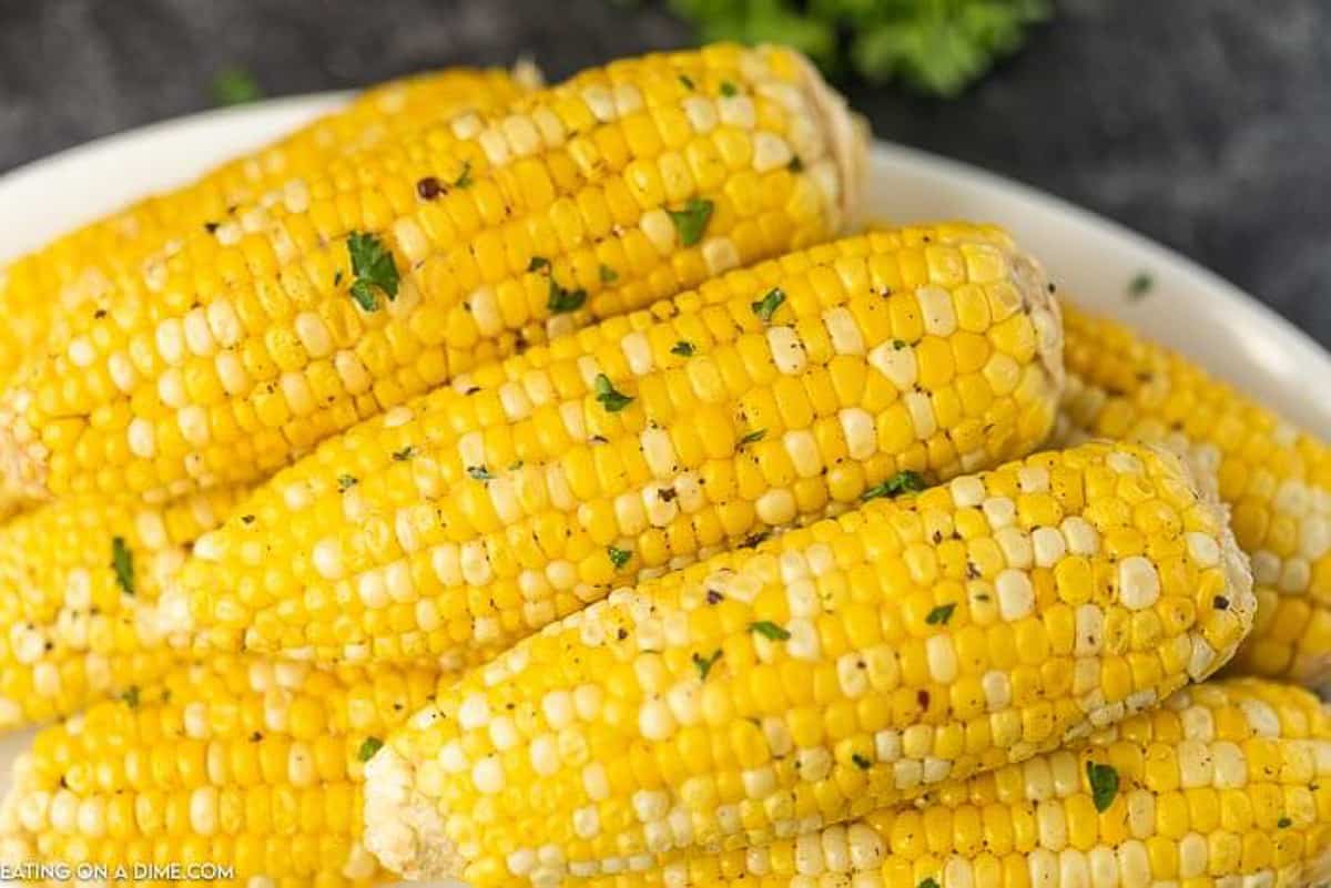 5 or 6 grilled corns on the cobs on a white plate topped with parsley and black pepper.