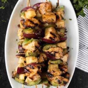 Grilled Hawaiian chicken kabobs recipe featuring chunks of chicken, green bell pepper, red onion, and pineapple on wooden skewers, served on a white oval plate.