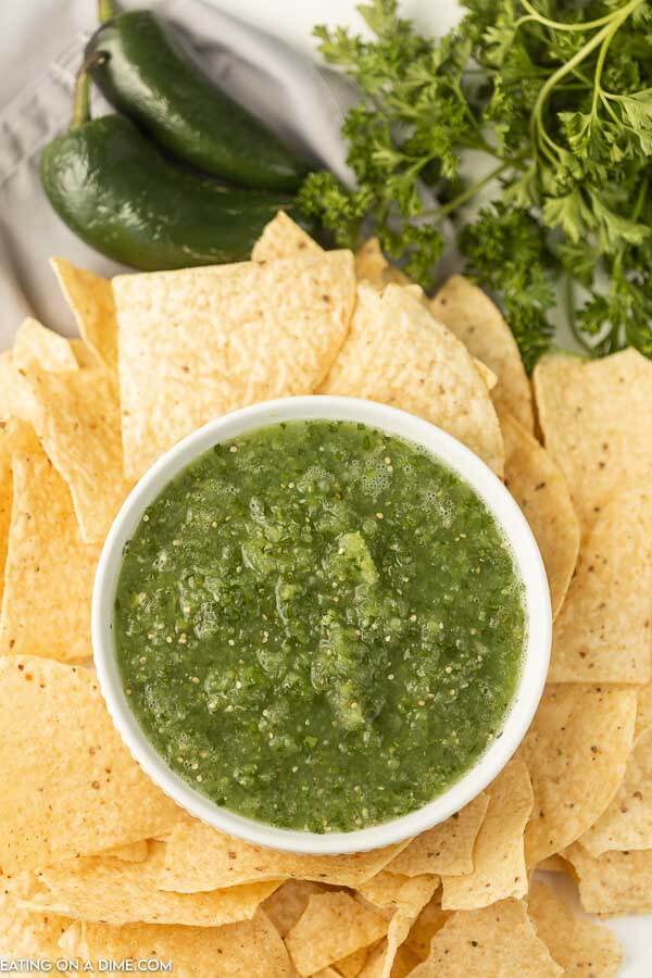 A white bowl of salsa verde surrounded by tortilla chips with jalapeños next to it 