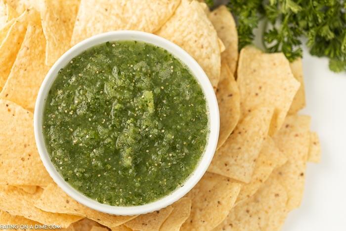 A white bowl of salsa verde surrounded by tortilla chips on a white countertop 