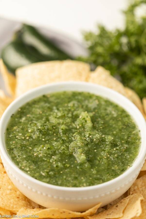 Close up of a white bowl of salsa verde surrounded by tortilla chips with jalapeños in the back ground 