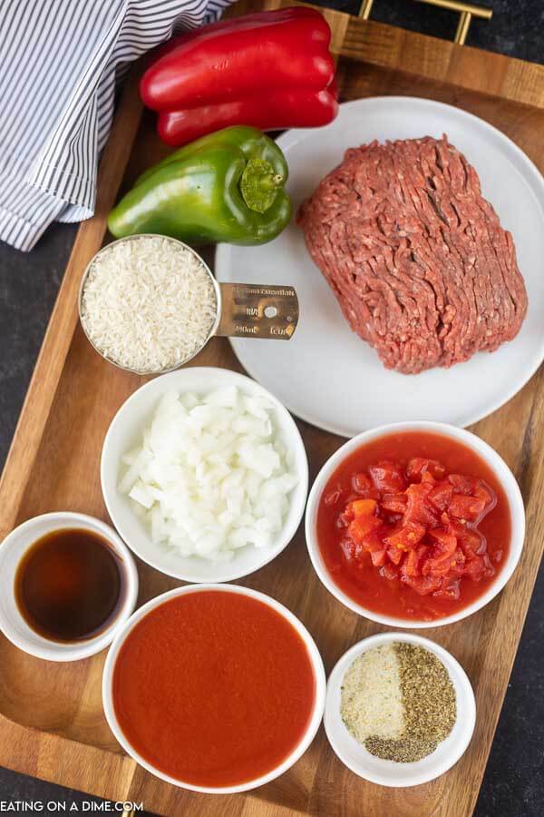Close up image of red and green pepper, rice in a measuring cup, uncooked ground beef, chopped onions, diced tomatoes, tomato sauce, worcestershire sauce, seasoning on a board. 