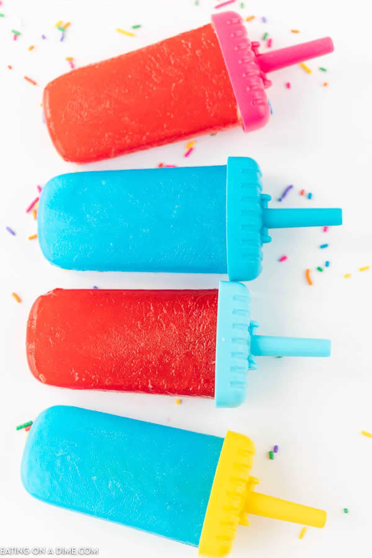 4 jello popsicles - 2 red and 2 blue on a white countertop with sprinkles scattered around them 