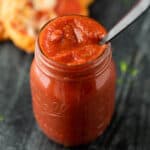 A glass jar filled with thick, red homemade pizza sauce recipe sits on a dark surface, with a spoon dipped into the sauce. Blurred food is visible in the background.
