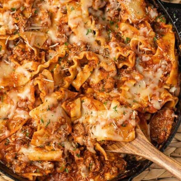 A close-up of a skillet lasagna recipe filled with cheesy, baked noodles, ground meat, tomato sauce, and melted cheese, with a wooden spoon lifting a serving.