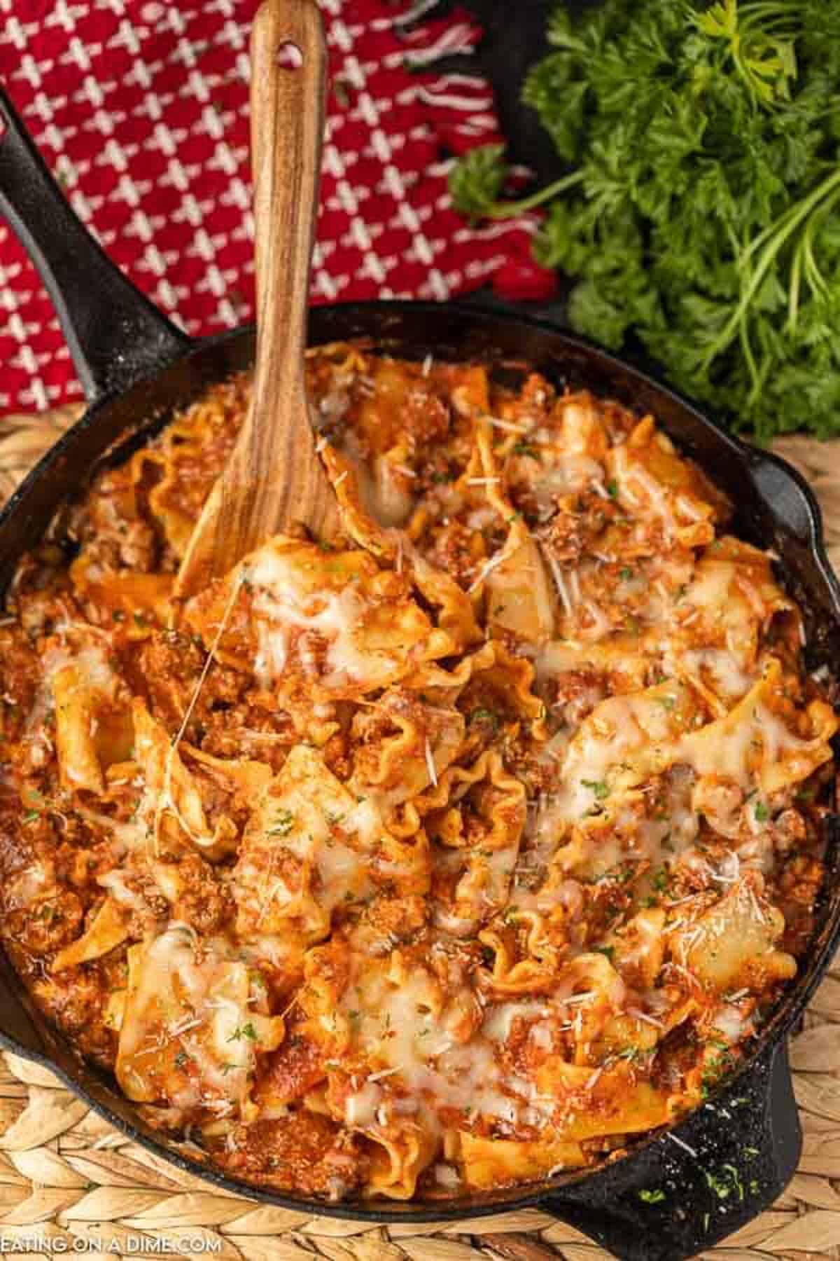 Lasagna in a cast iron skillet with a wooden spoon in it with a red linen and fresh parsley in the background. 