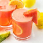 Two glasses of pink Homemade Watermelon Lemonade garnished with lemon slices and a wedge of watermelon, with extra lemon and watermelon pieces in the background on a white surface.