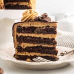 A slice of Chocolate Peanut Butter Cake with peanut butter frosting, topped with a swirl of frosting and pieces of peanut butter cups, sits on a white plate with a fork.