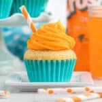 An Orange Soda Cupcake in a bright blue wrapper, topped with swirled orange frosting and an orange-striped straw, sits on a white plate. Orange soda and matching straws are visible in the background.