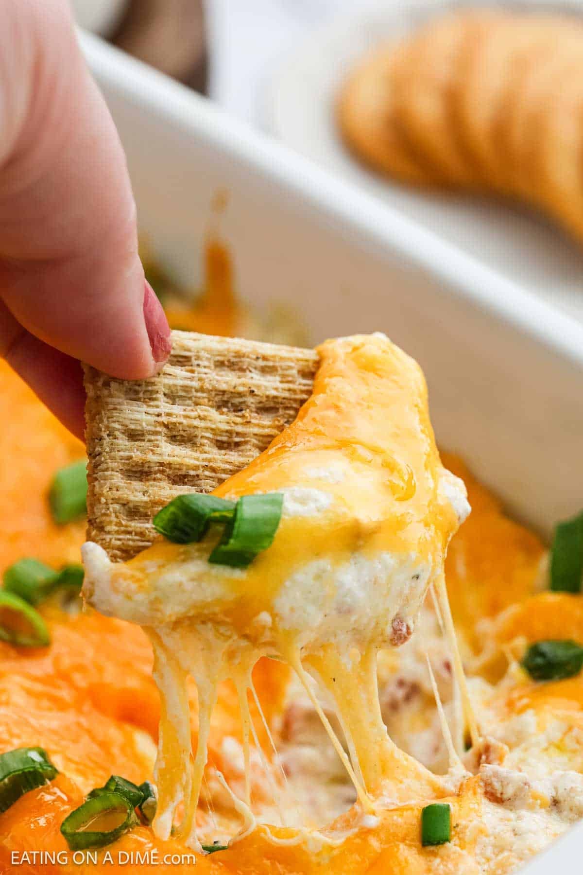 A hand dips a square cracker into a cheesy, baked Crack Dip topped with melted cheddar and chopped green onions, with gooey cheese stretching to the cracker. Crackers are visible in the background.